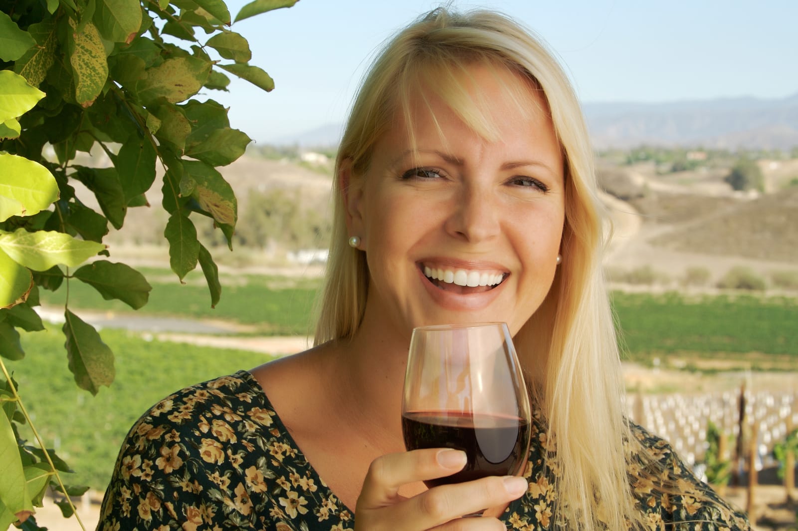 Smiling Blonde Woman in a Floral Dress Holding a Glass of Red Wine Outdoors in a Vineyard.
