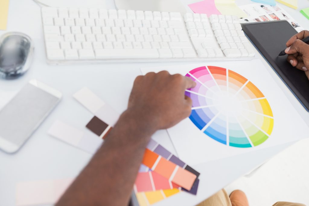 Designer’s Hands Work with a Color Wheel and Swatches on a White Desk, with a Keyboard and Drawing Tablet Nearby.