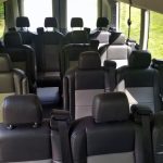 Interior of a Passenger Van with Multiple Rows of Black Leather Seats and Seat Belts; Green Outdoors Visible Through Windows.